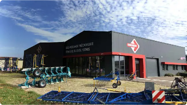 Outdoors view of a black farm equipment workshop with a red entrance, displaying blue and teal cultivators and other machinery in the yard.