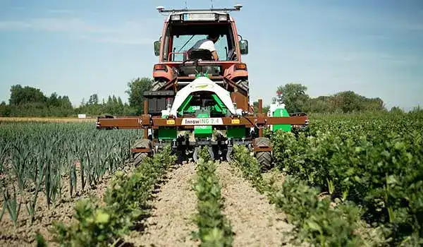 Red farm tractor towing a machine carrying a white-and-green jet ski along a dirt path between crop rows under a blue sky.