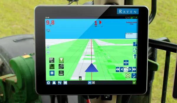 In-cab Raven precision farming display showing automated steering line on a green field, blue sky, and control icons.