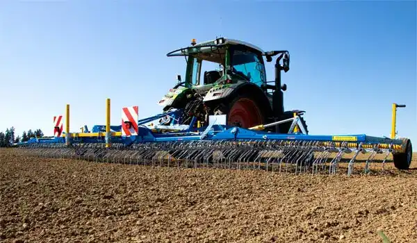 Green tractor towing a large blue cultivator across a freshly tilled field under a clear blue sky.
